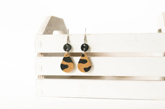 A pair of tan leopard suede leather earrings displayed on a white wooden earring stand.